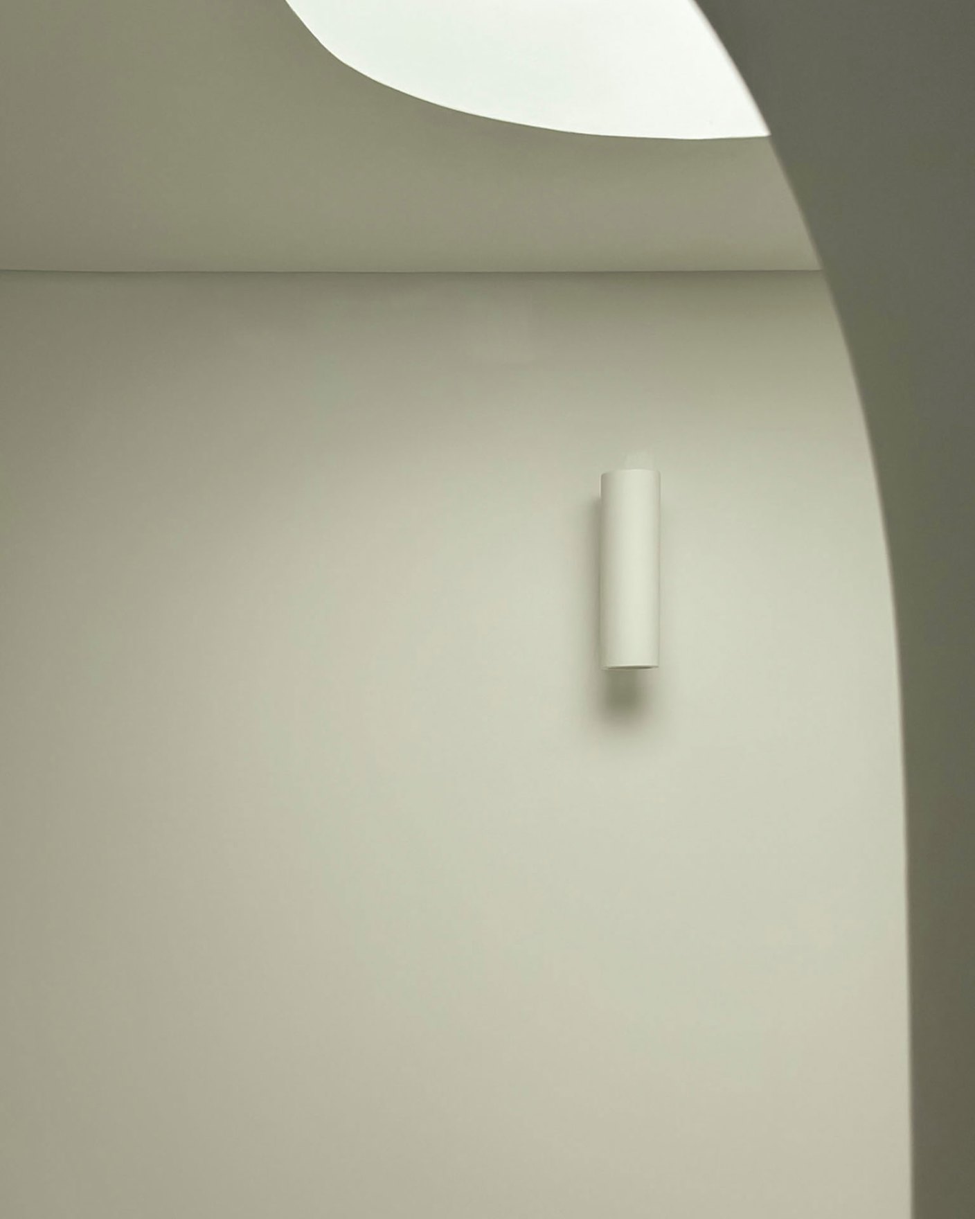 Detail of a curved plaster wall with a slim cylindrical wall light and a soft skylight above