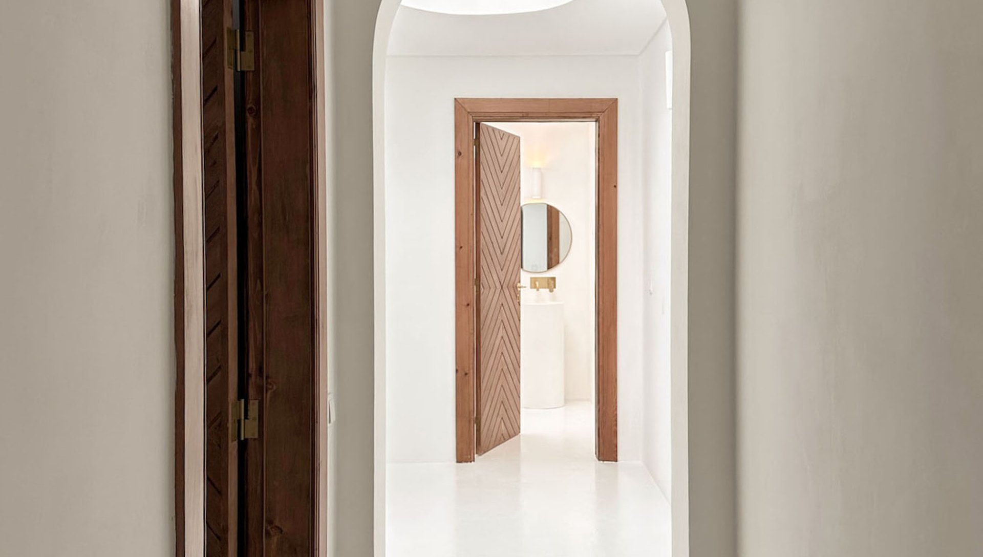 Whitewashed corridor framed by an arched opening, with a round skylight beyond and a solid timber door