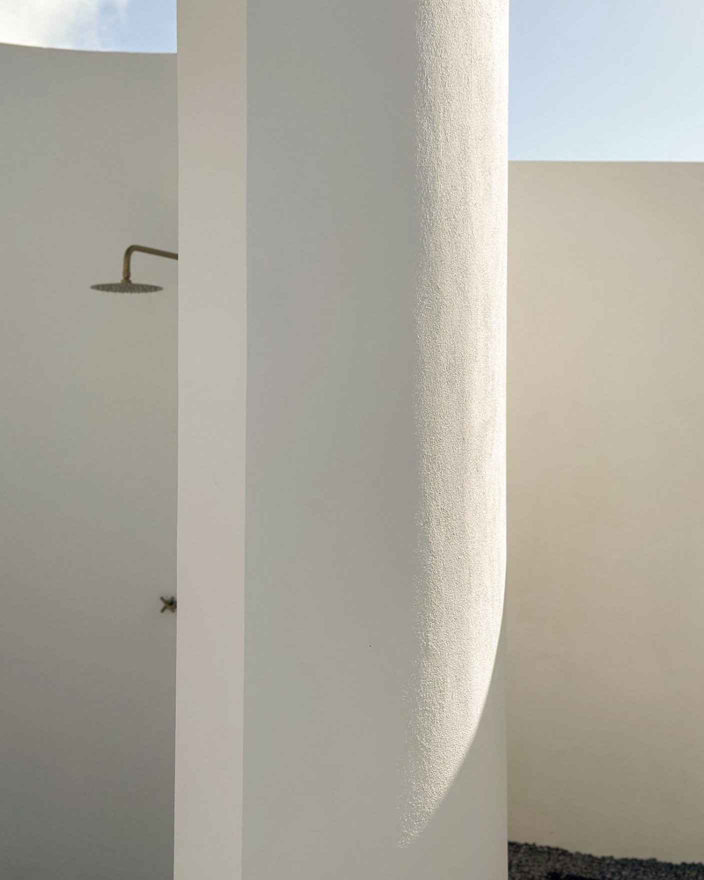 Curved white plaster wall in raking sunlight beside an outdoor brass shower